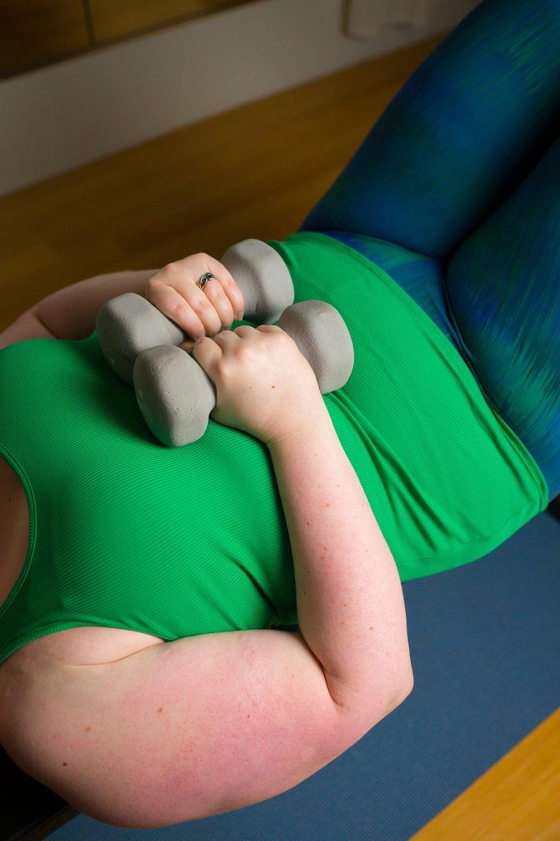 A person with a green tank top and blue and green athletic pants laying on their back, holding 2 small grey dumbbells together over their stomach.